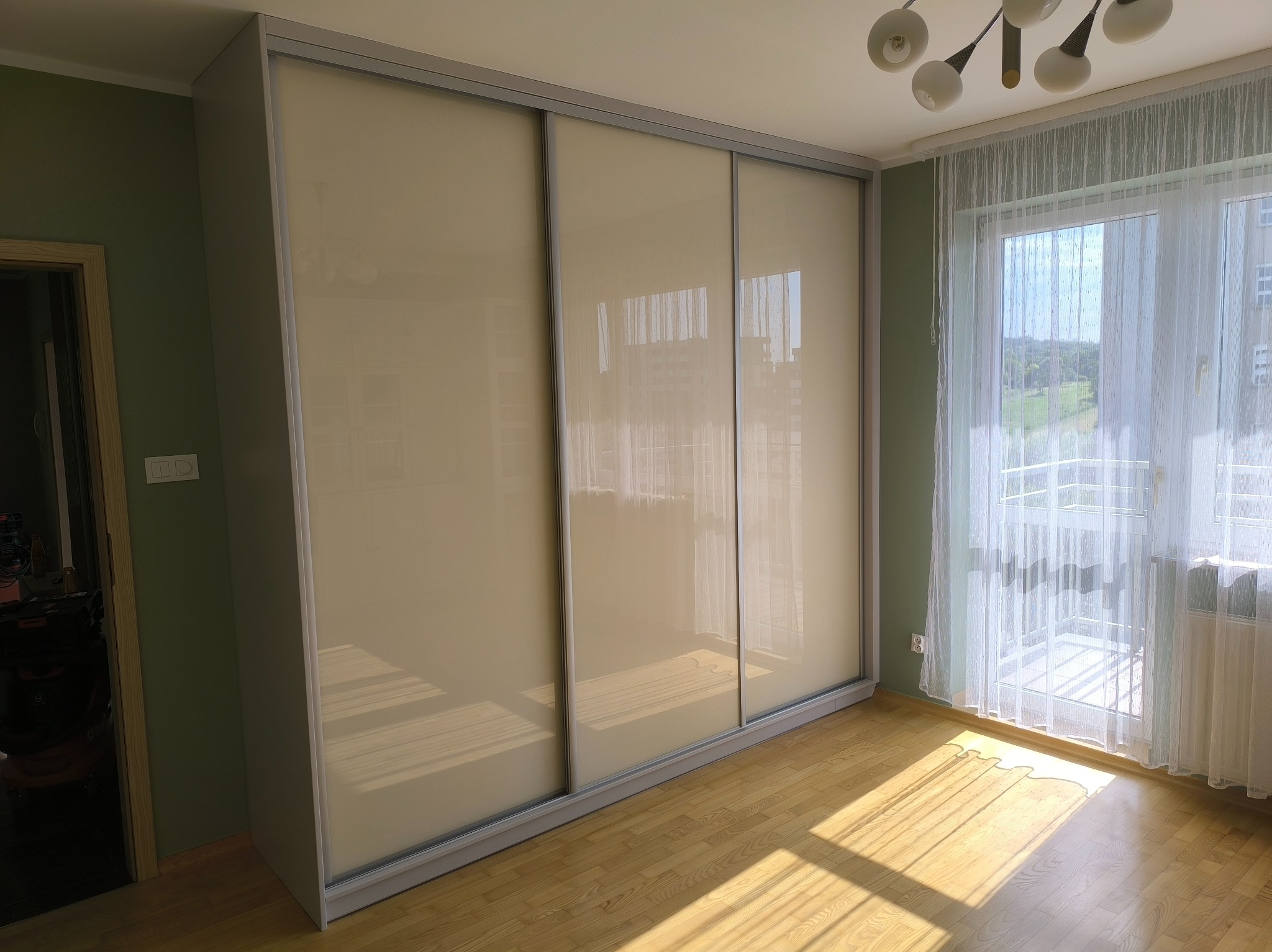 Jasna szafa wnękowa z przesuwnymi drzwiami, wkomponowana w pokój z parkietem i oknem balkonowym, widziana w świetle dziennym.