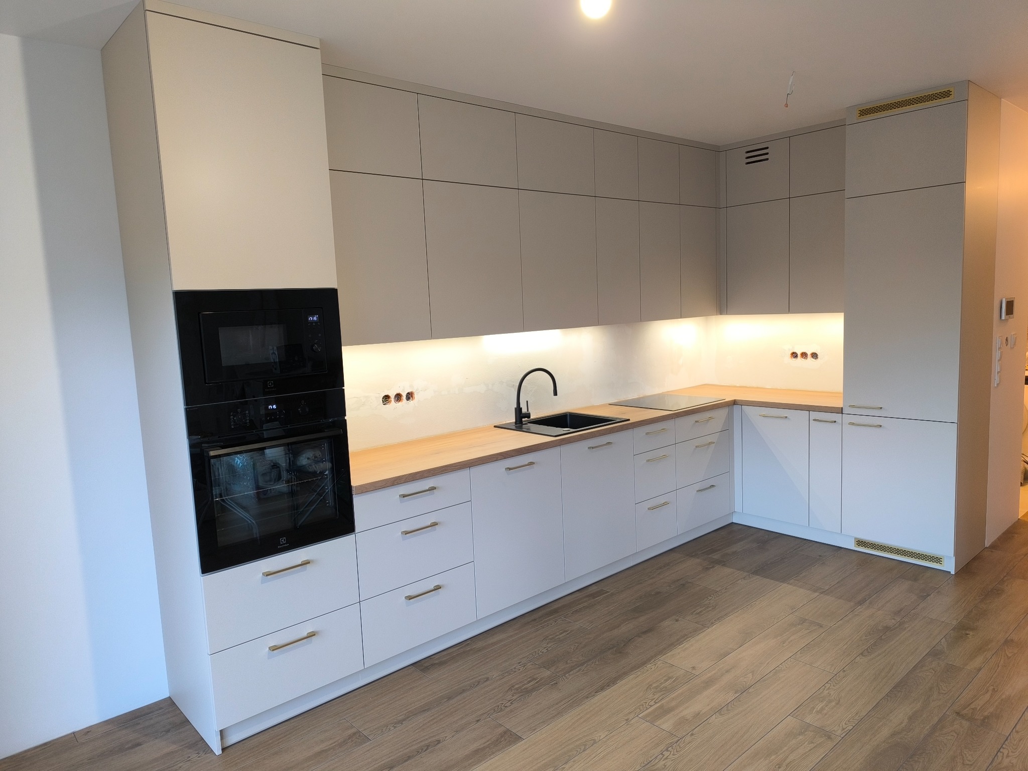 Jasna, nowoczesna kuchnia z drewnianym blatem i czarną armaturą. Zabudowa pod sufit z piekarnikiem i mikrofalówką. Parkiet na podłodze. Minimalistyczny design.