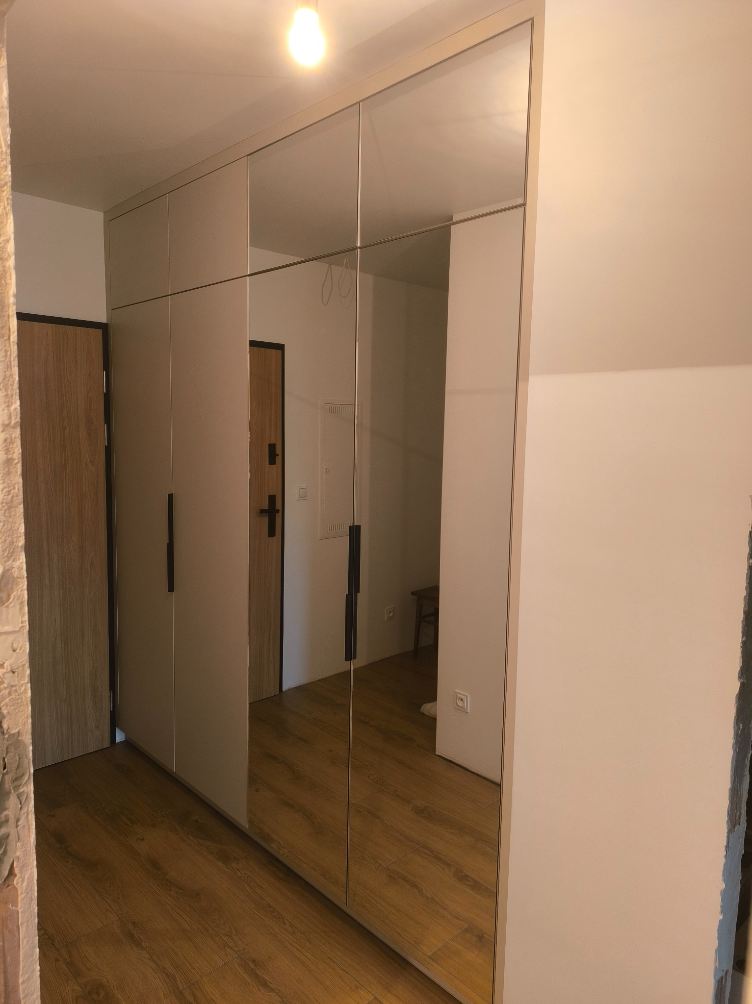 Szafa wnękowa z lustrzanymi drzwiami przesuwnymi, z minimalistycznymi, czarnymi uchwytami, w przedpokoju z drewnianą podłogą i widocznymi fragmentami ściany w trakcie remontu.