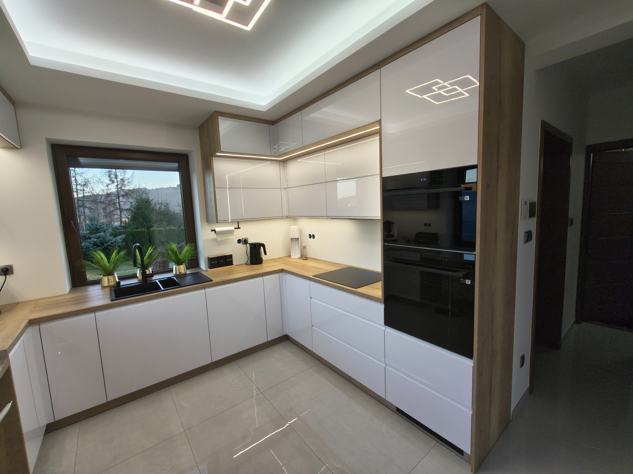 Nowoczesna kuchnia w bieli i drewnie z oświetleniem LED, czarnym zlewem i sprzętem AGD. Widok na okno z zielenią i fragment korytarza.