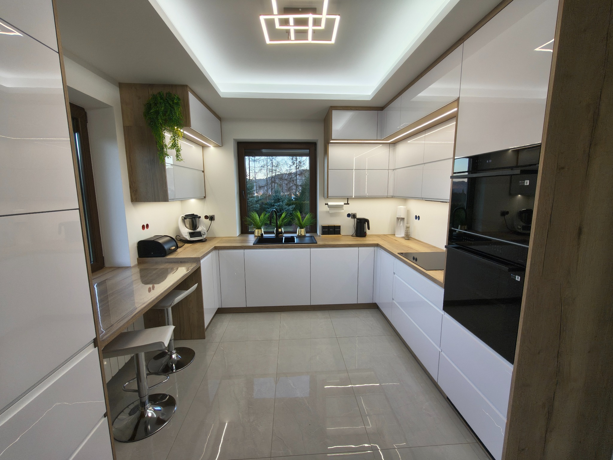 Nowoczesna kuchnia z białymi, lakierowanymi frontami i drewnianymi akcentami. Uwagę przykuwa designerska lampa sufitowa i rośliny doniczkowe przy oknie.