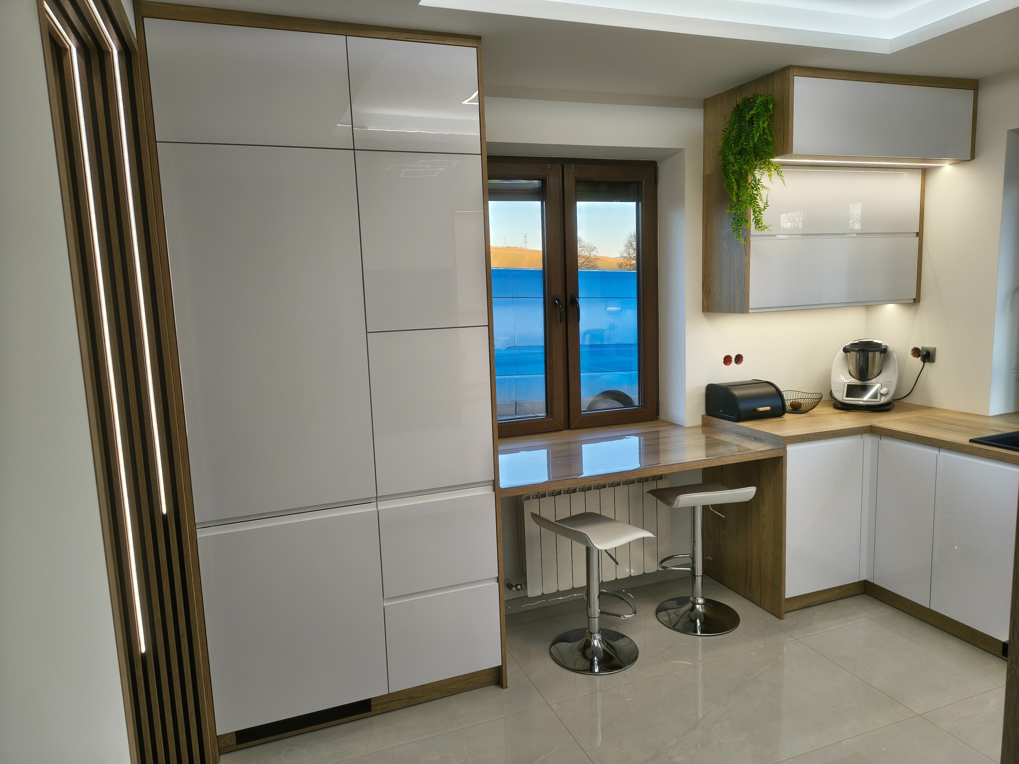 Nowoczesna kuchnia z białymi, lakierowanymi frontami, drewnianymi akcentami i oświetleniem LED. Blat roboczy zintegrowany z parapetem, dwa hokery. Widok na zewnątrz.