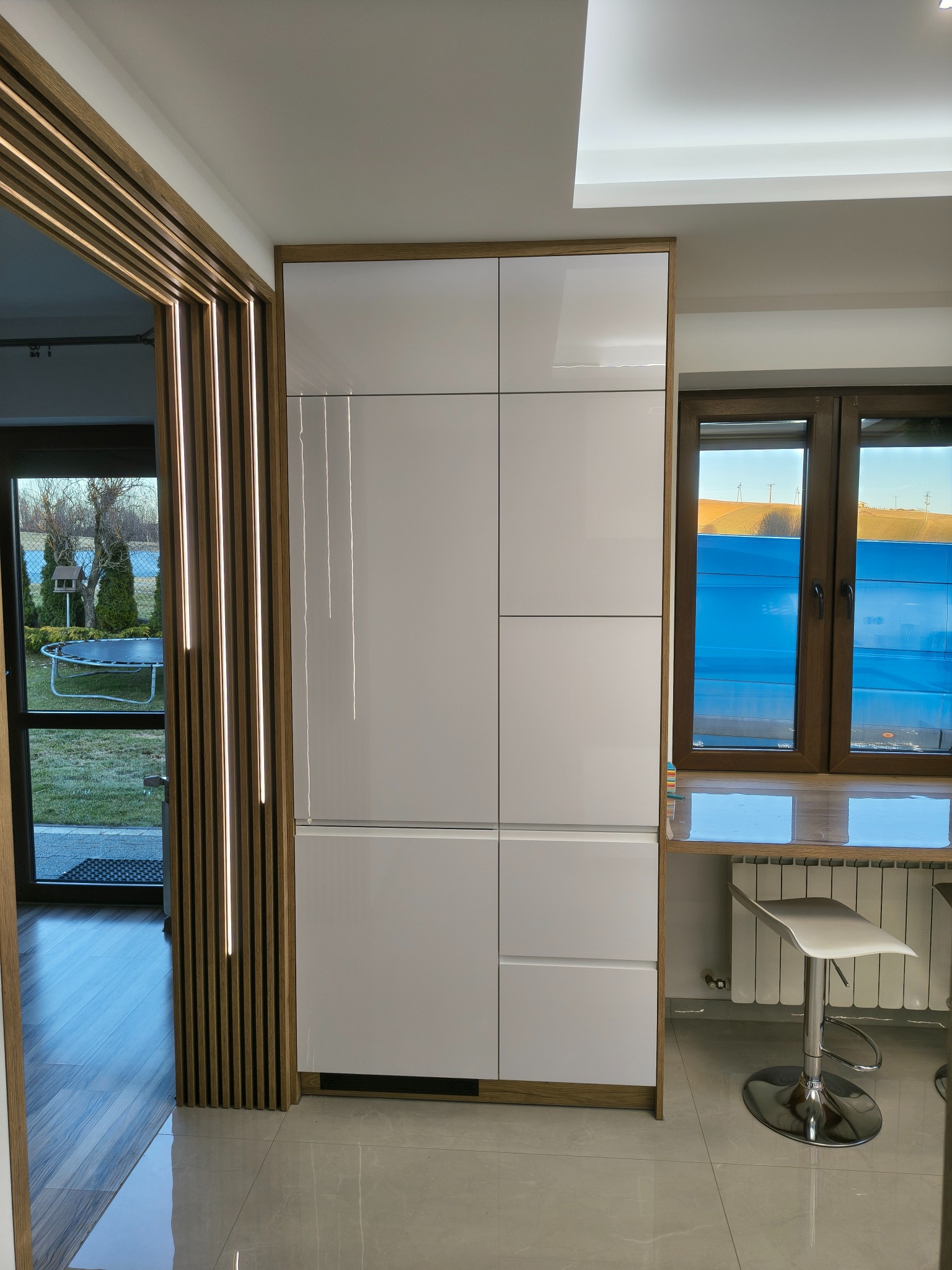 Biała, lakierowana szafa w zabudowie z drewnianymi akcentami, w nowoczesnej kuchni. Widoczne okno z blatem i krzesłem barowym. Dekoracyjne lamele przy wejściu.