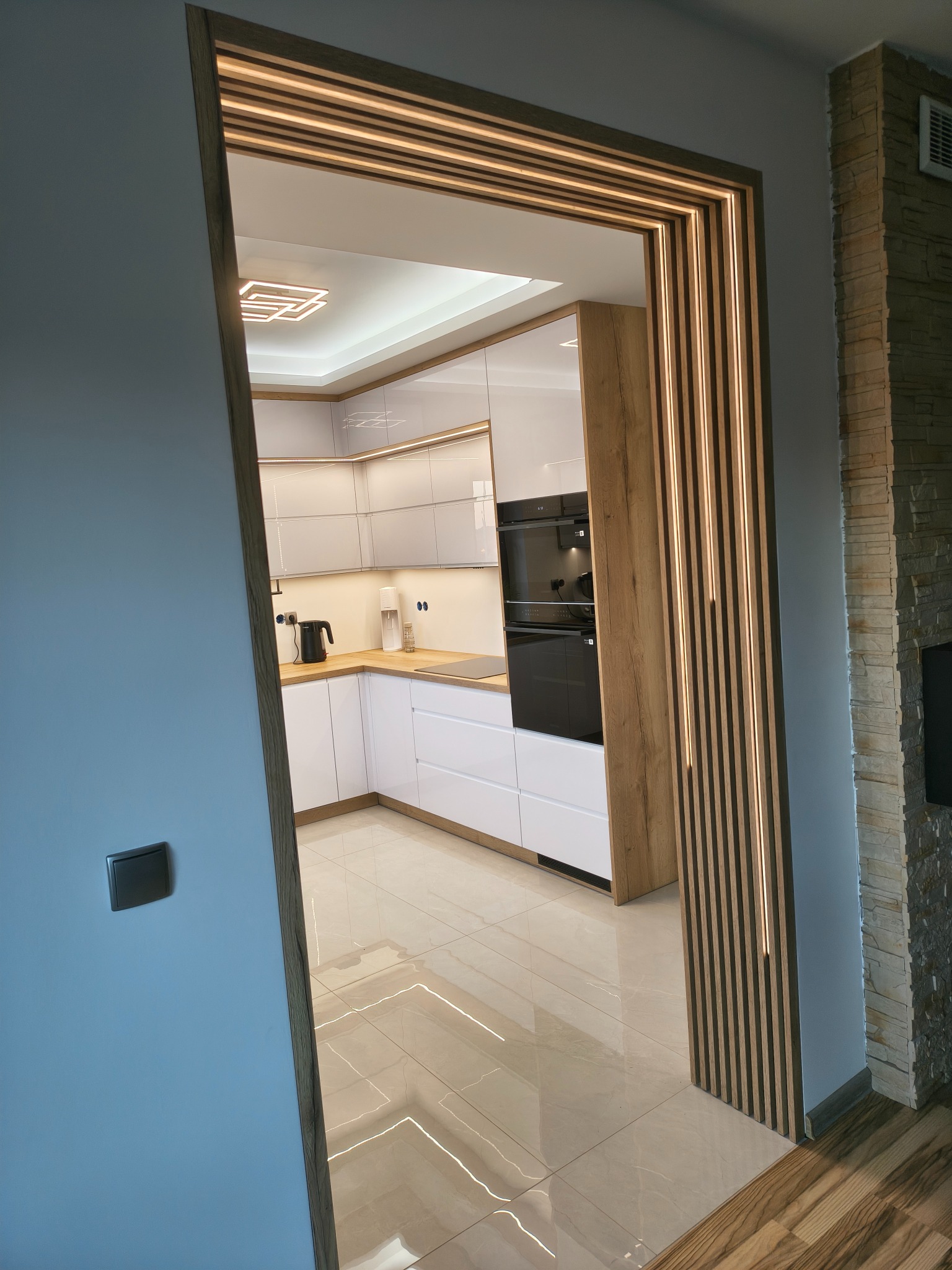Nowoczesna kuchnia z białymi frontami, drewnianym blatem i oświetleniem LED, widziana przez ozdobne przejście z drewnianymi lamelami i podświetleniem.