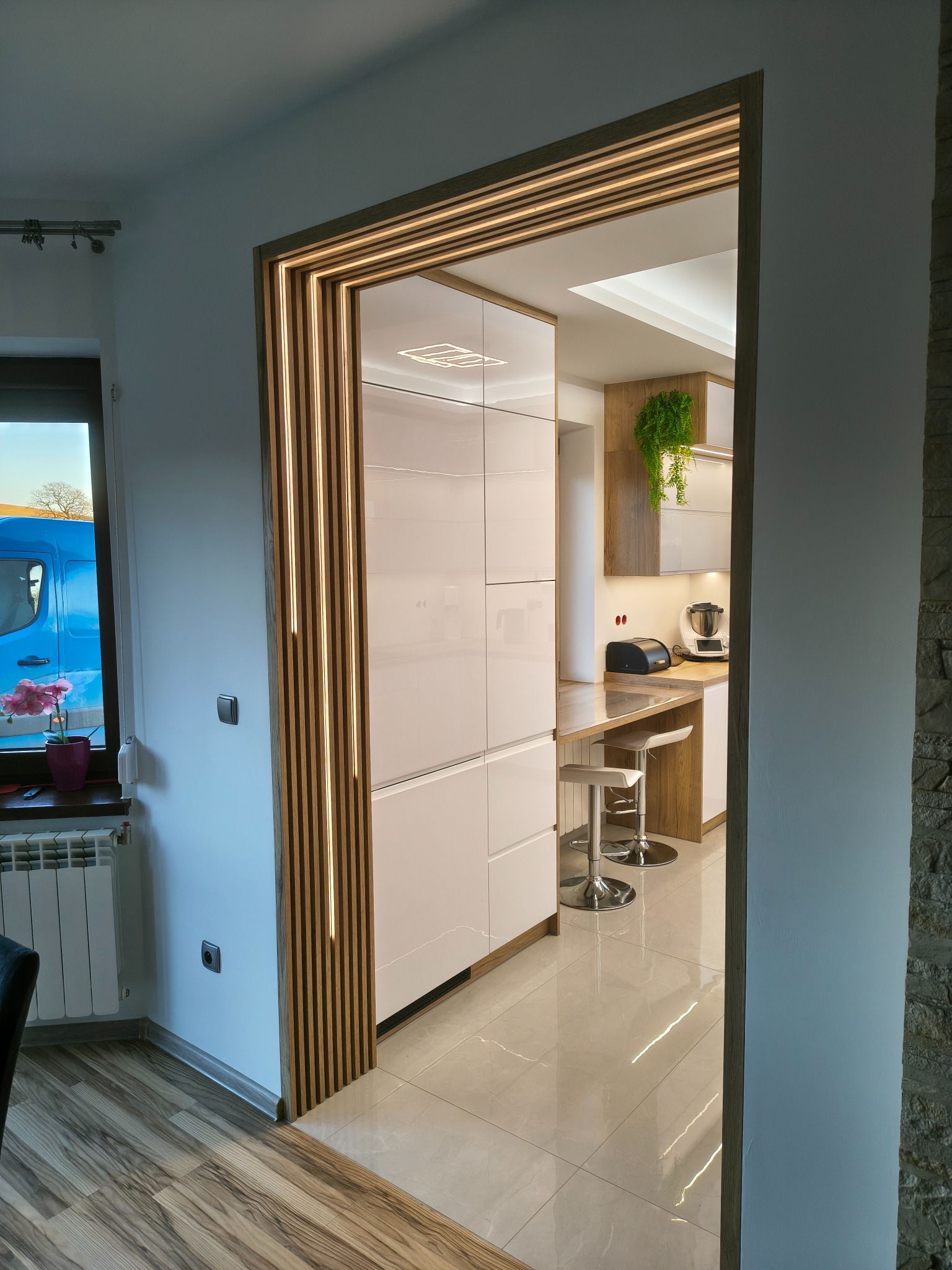 Nowoczesna kuchnia z białymi frontami, widoczna przez drewnianą ramę z oświetleniem LED. Blat z hokerami, rośliny doniczkowe. Jasne płytki podłogowe.