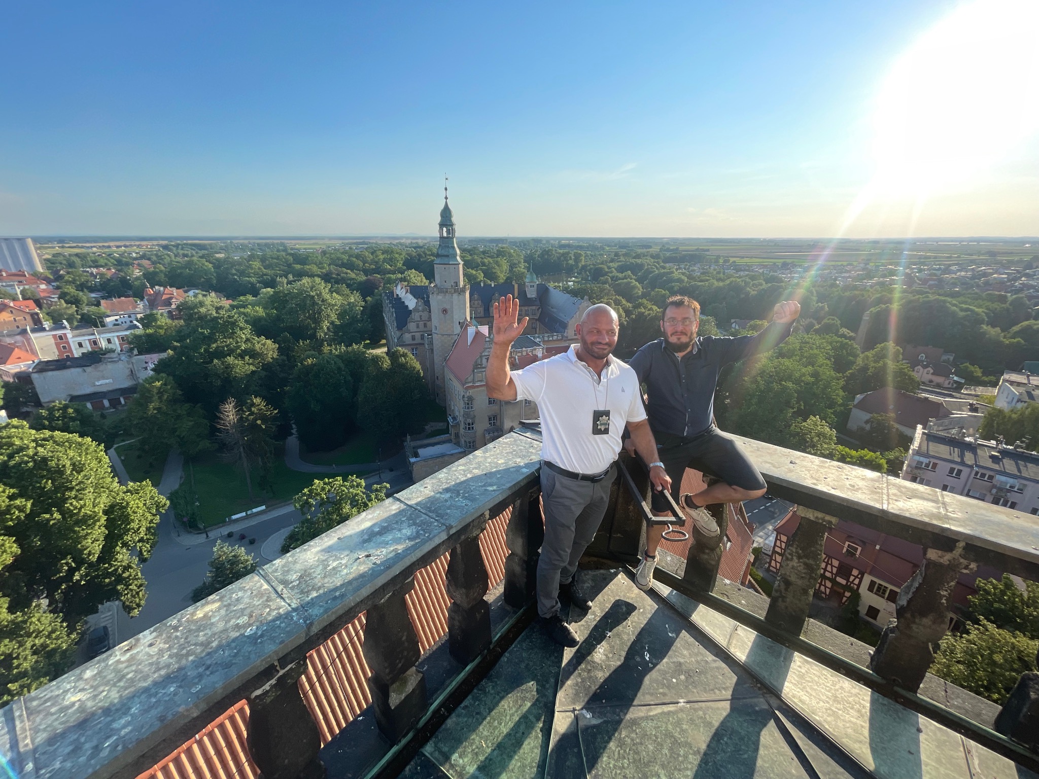Dwóch mężczyzn na szczycie wieży zamkowej, jeden w białej koszuli z identyfikatorem, drugi w ciemnej koszuli, obaj machają rękami, panorama miasta w tle.