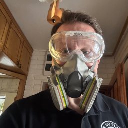 Pan do Plam Kompleksowo dla Czystości Artur Janiec - Osoba w masce i okularach ochronnych, ubrana w czarną koszulkę z logo firmy sprzątającej, spogląda w dół z perspektywy sufitu, w tle widoczne fragmenty mebli i lampy.