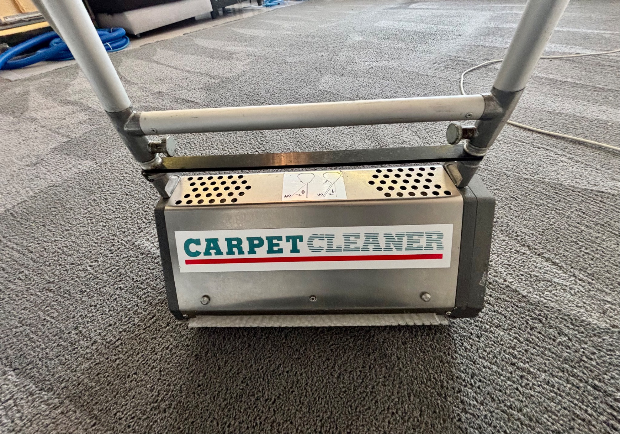 Profesjonalna maszyna do czyszczenia dywanów z napisem 'CARPET CLEANER' na tle szarego, wyczyszczonego dywanu.