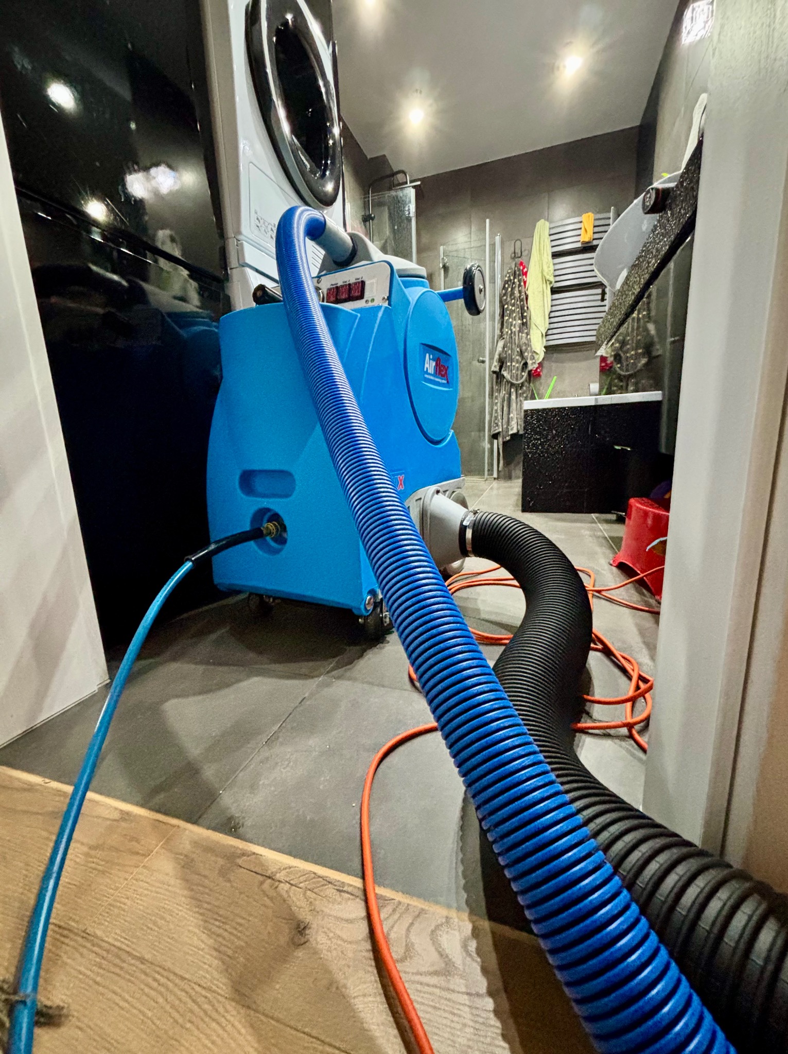 Niebieskie urządzenie AirFlex z podłączonymi wężami w łazience, prawdopodobnie do czyszczenia. W tle pralka i wyposażenie łazienki.