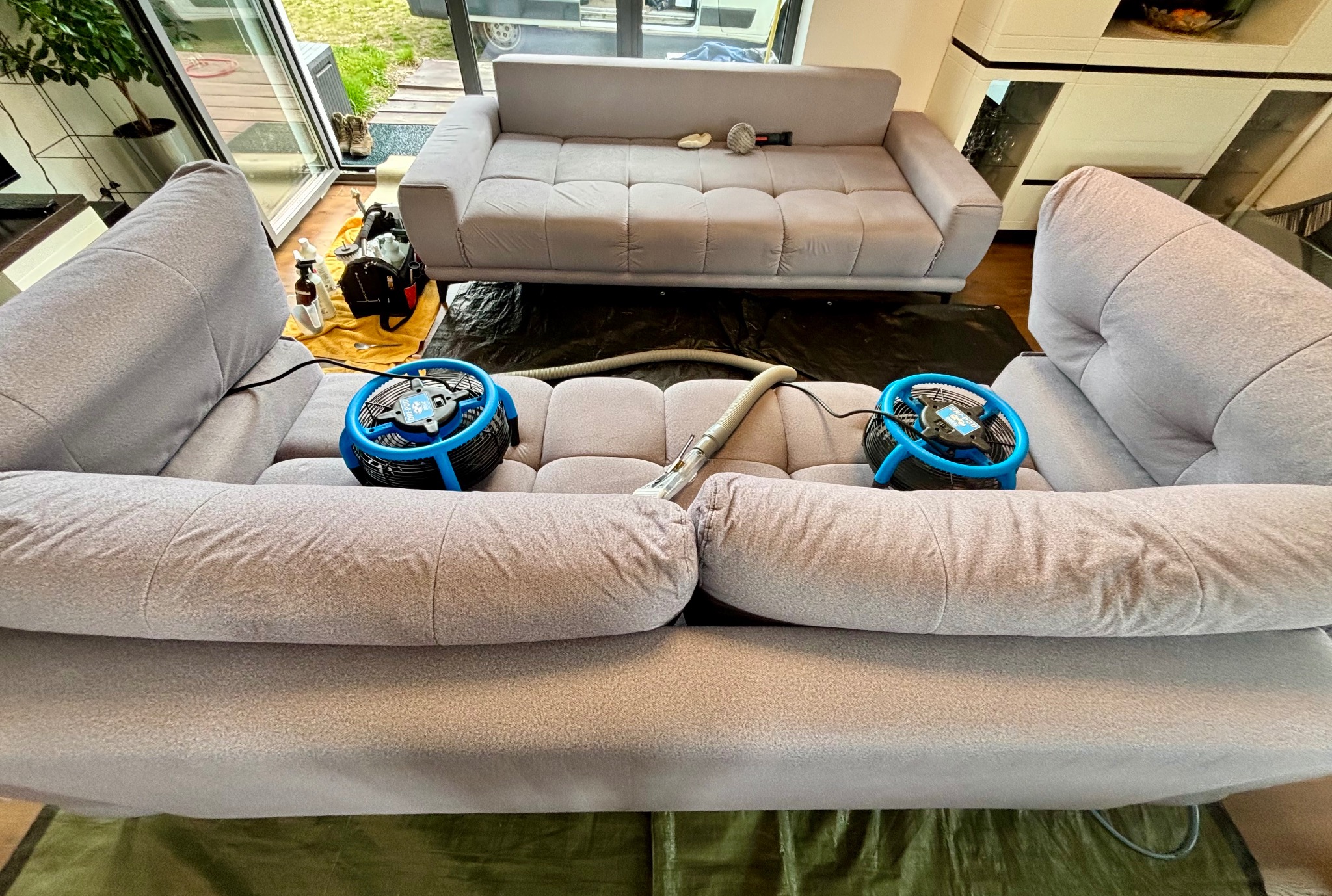 Szara kanapa w trakcie czyszczenia z użyciem odkurzacza piorącego i wentylatorów. Druga kanapa w tle, zabezpieczona folią. Widoczne narzędzia i środki czystości.