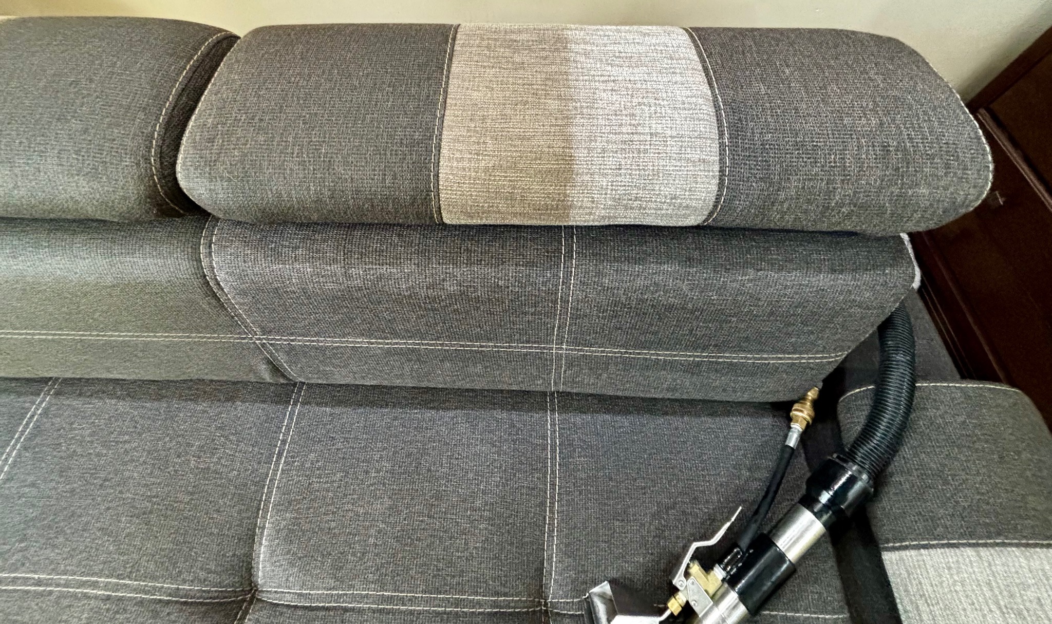 Szara kanapa z widocznym jasnym pasem na oparciu, na której leży dysza do prania tapicerki z czarnym wężem. Widoczne szwy i faktura materiału.