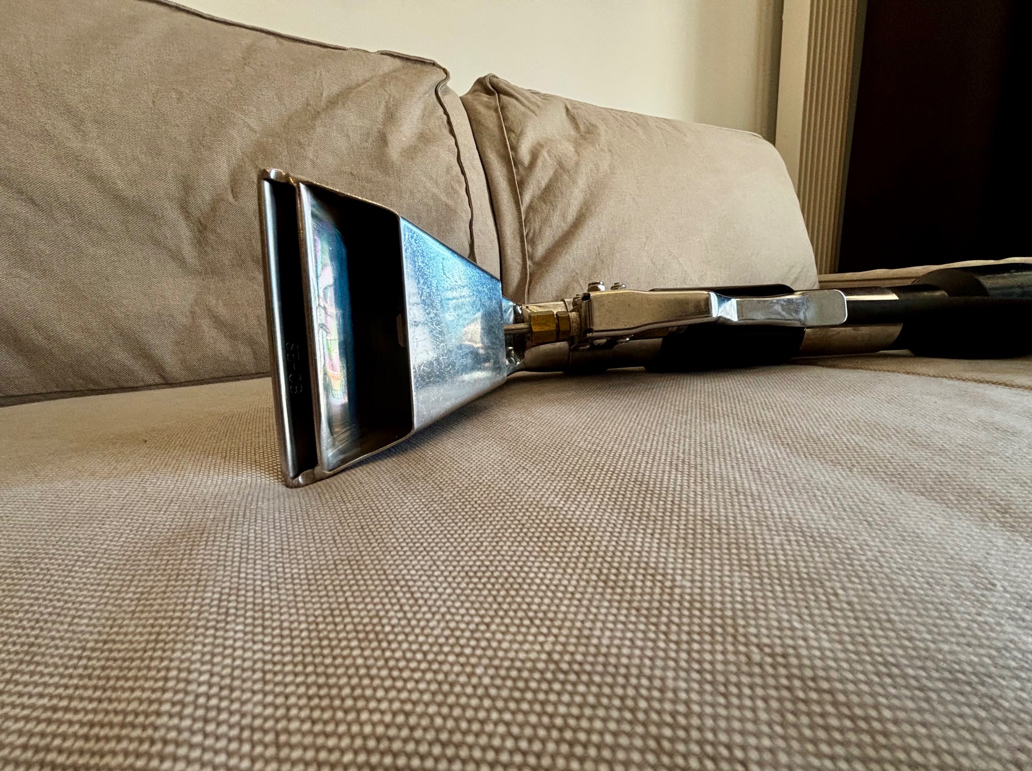 Dysza do prania tapicerki na beżowej kanapie. Metalowa końcówka i czarny wąż. Widoczne detale tkaniny. Sprzęt do czyszczenia.