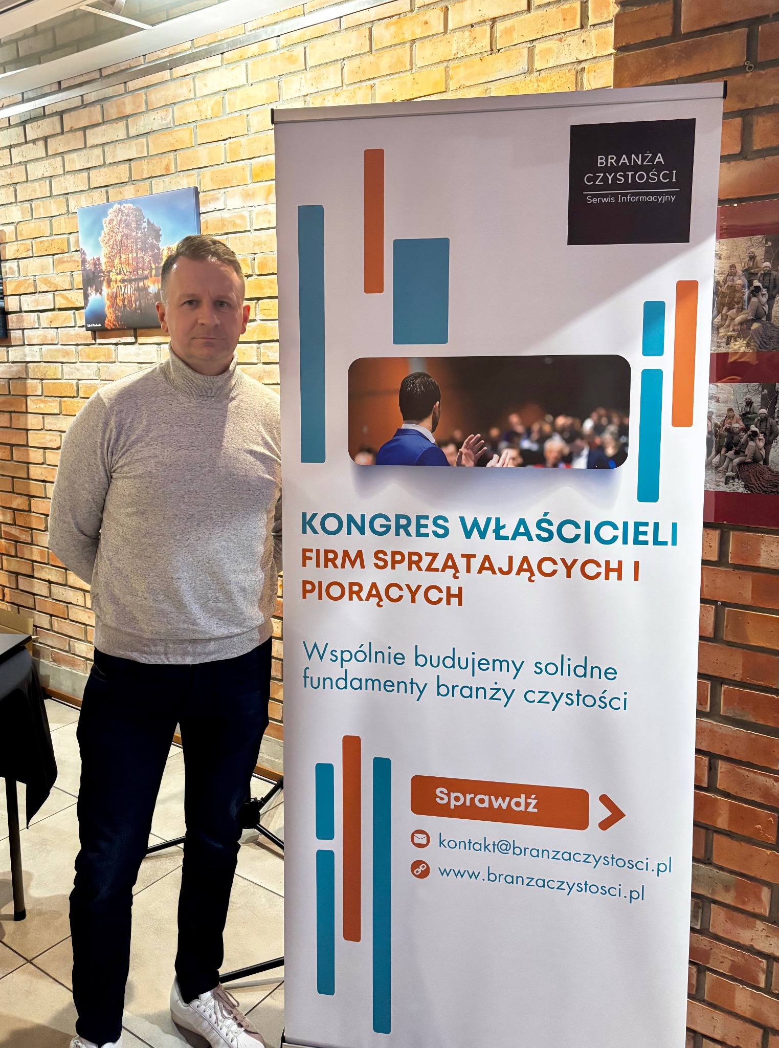 Mężczyzna w szarym golfie pozuje obok roll-upu Kongresu Właścicieli Firm Sprzątających i Piorących na tle ceglanej ściany. Widoczny adres e-mail i strona www.