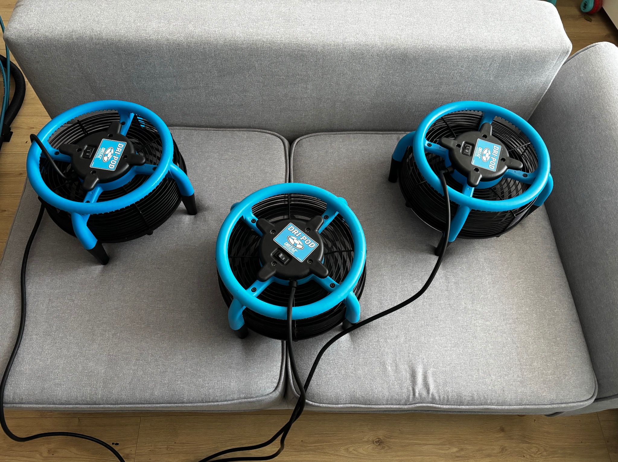 Profesjonalne suszarki do tapicerki DRI-POD na szarej kanapie. Urządzenia z niebieskimi akcentami, czarne kable. Widok z góry na proces suszenia po czyszczeniu.