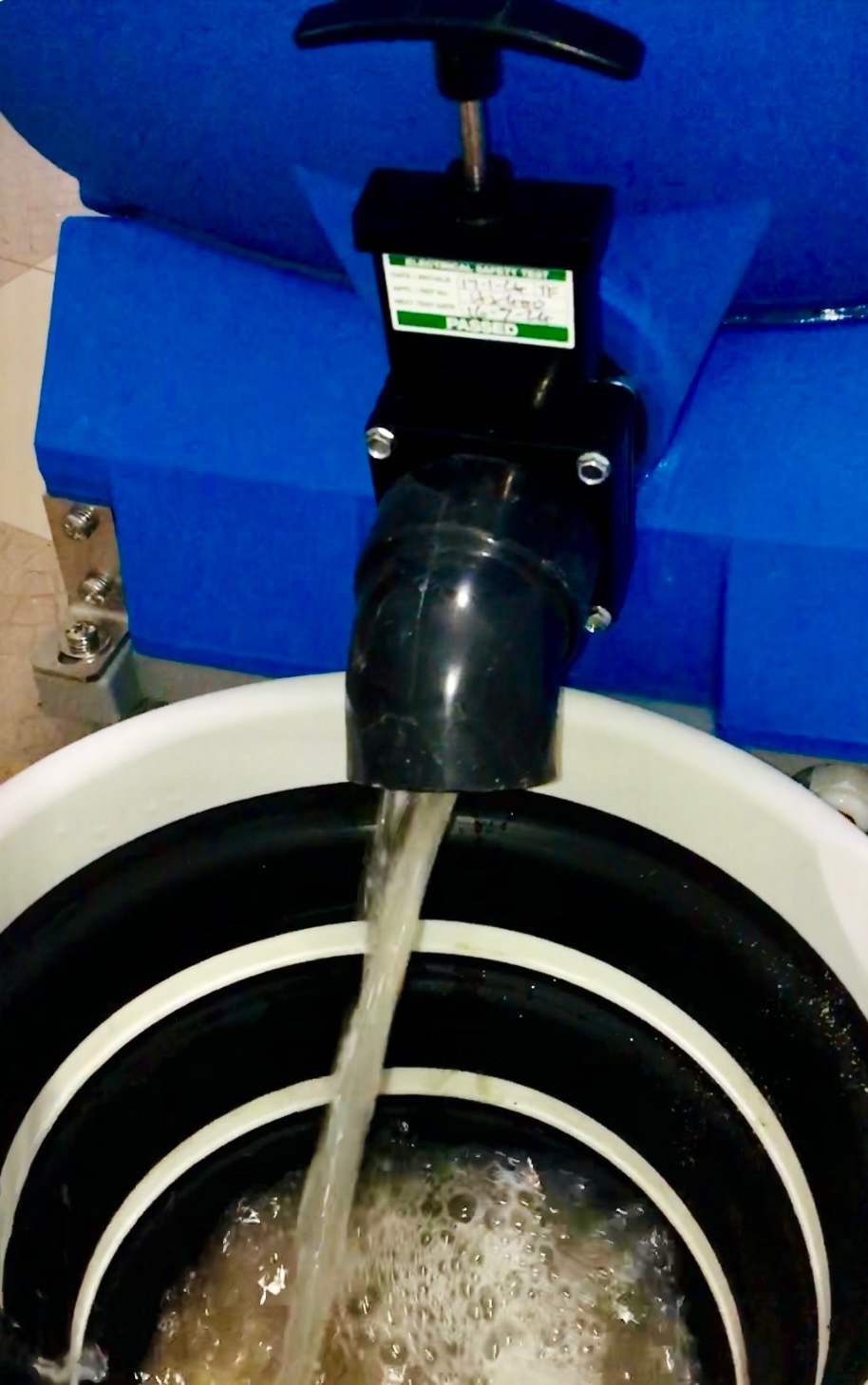 Woda wypływająca z czarnego węża do białego wiadra z czarno-białymi okręgami, w tle niebieski element urządzenia z zaworem i naklejką 'Passed'.