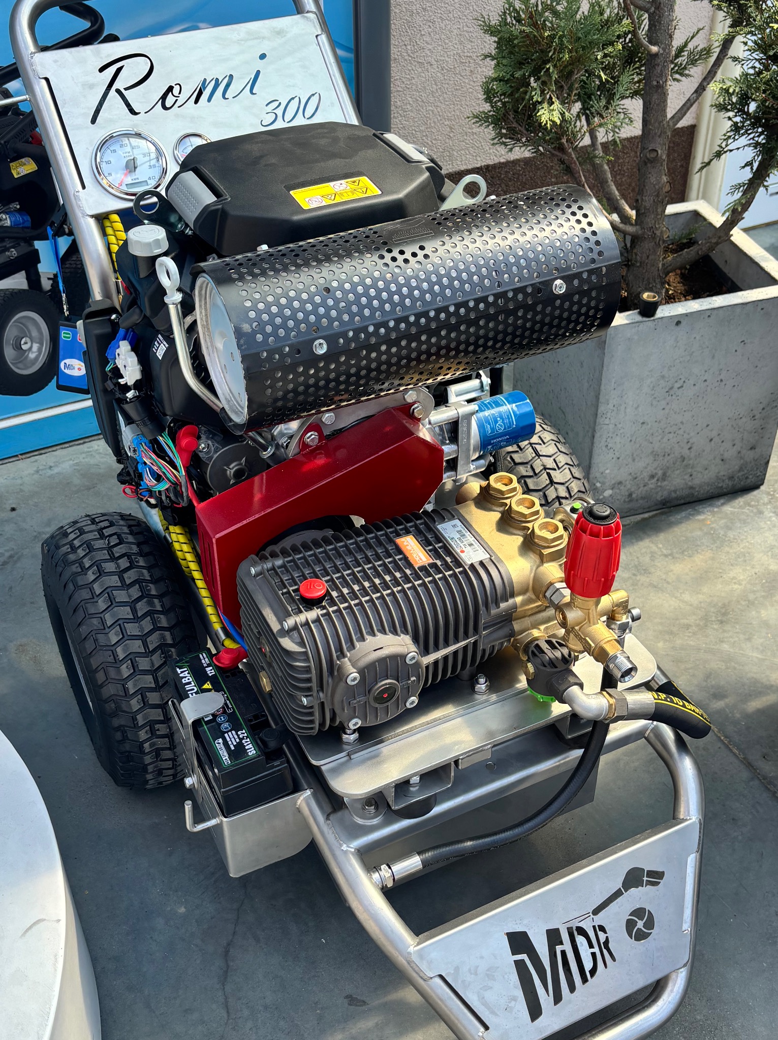 Profesjonalna myjka ciśnieniowa Romi 300 z silnikiem spalinowym i logotypem MDR na stalowej ramie, stojąca na szarym podłożu, z drzewkiem w donicy w tle.