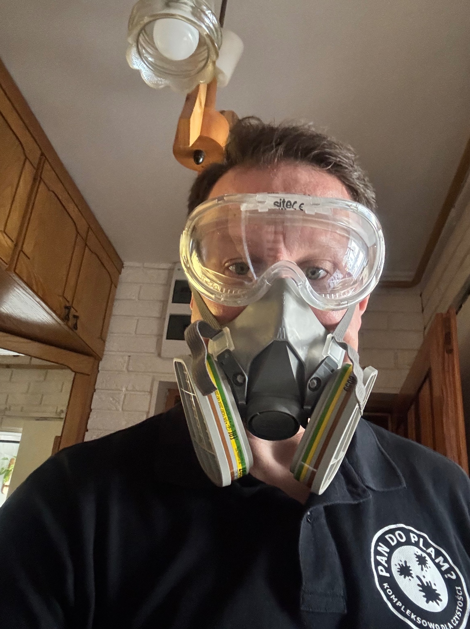 Osoba w masce i okularach ochronnych, ubrana w czarną koszulkę z logo firmy sprzątającej, spogląda w dół z perspektywy sufitu, w tle widoczne fragmenty mebli i lampy.