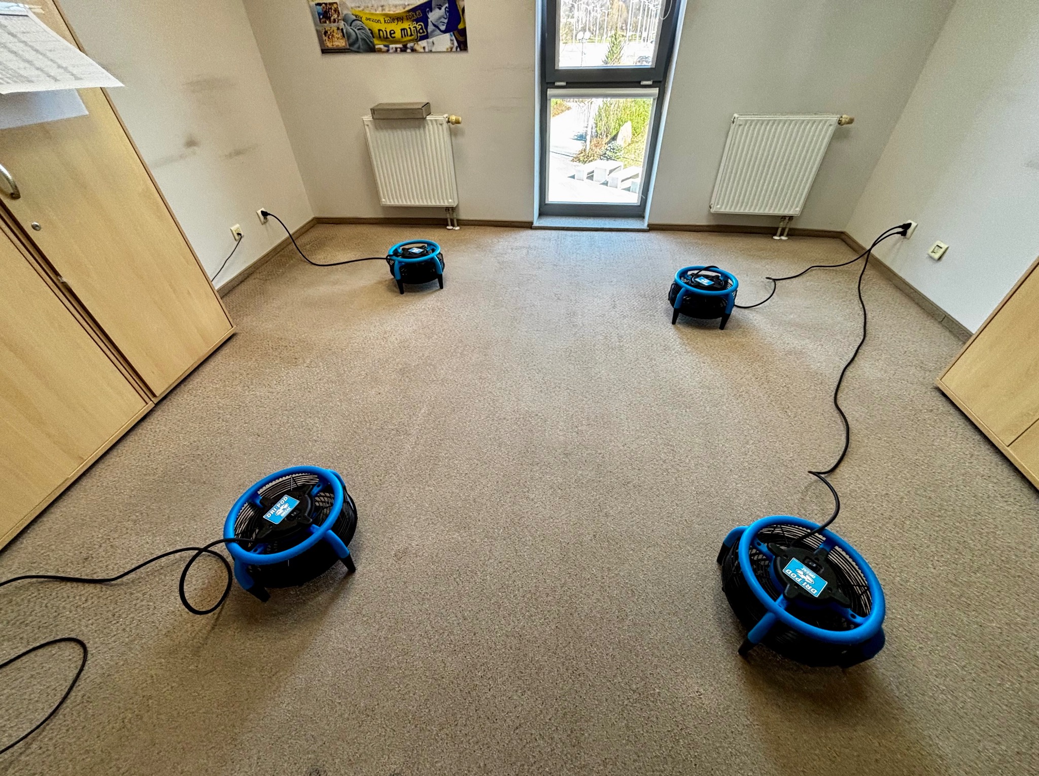 Cztery niebieskie wentylatory osuszające DRI-POD na beżowej wykładzinie w pokoju z oknem, szafą i grzejnikiem. Urządzenia podłączone do gniazdek elektrycznych.