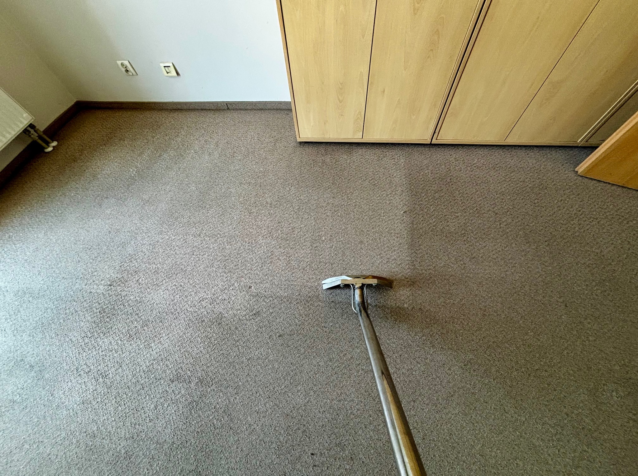 Pranie dywanu w biurze: fragment wyczyszczonego dywanu z widoczną różnicą w kolorze i urządzenie do prania dywanów z metalową rączką.