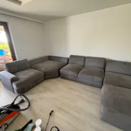 Szara modułowa sofa narożna w trakcie czyszczenia, widoczny odkurzacz piorący i akcesoria na jasnej podłodze.