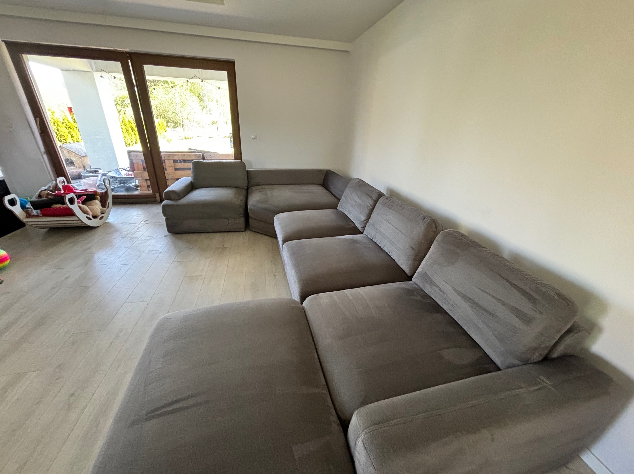 Szara, modułowa sofa narożna w jasnym salonie z widokiem na ogród przez duże okno. Na podłodze widoczne ślady użytkowania.