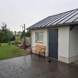 USŁUGI BUDOWLANE KAMIL KIEŁBOWICZ BARON-BUD - Biały budynek gospodarczy z szarą metalową dachówką, ciemnoszarymi drzwiami, małym oknem i drewnianą ławką przed wejściem, na mokrym brukowanym podjeździe. W tle trawnik, drzewa i element małej...