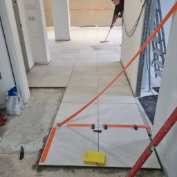 USŁUGI BUDOWLANE BARON-BUD - Układanie jasnych płytek podłogowych w korytarzu, widoczne narzędzia glazurnicze, poziomica, gąbka oraz osoba myjąca podłogę w tle.