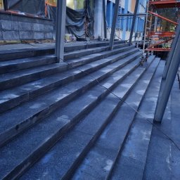 USŁUGI BUDOWLANE KAMIL KIEŁBOWICZ BARON-BUD - Granitowe schody zewnętrzne w trakcie montażu, widoczne rusztowania i elementy konstrukcyjne budynku, ciemne wykończenie.