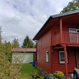 USŁUGI BUDOWLANE BARON-BUD - Czerwony, drewniany dom z balkonem w trakcie malowania. Widoczne pędzle, farby i drabina. Obok zielony budynek gospodarczy i osoba pracująca przy elewacji.