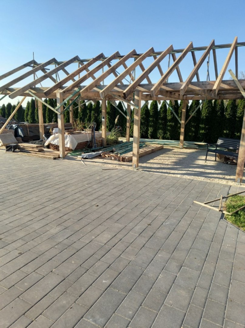 Konstrukcja drewniana zadaszenia tarasu w trakcie budowy, widoczne elementy drewniane, podłoże z kostki brukowej i rząd tuj w tle.