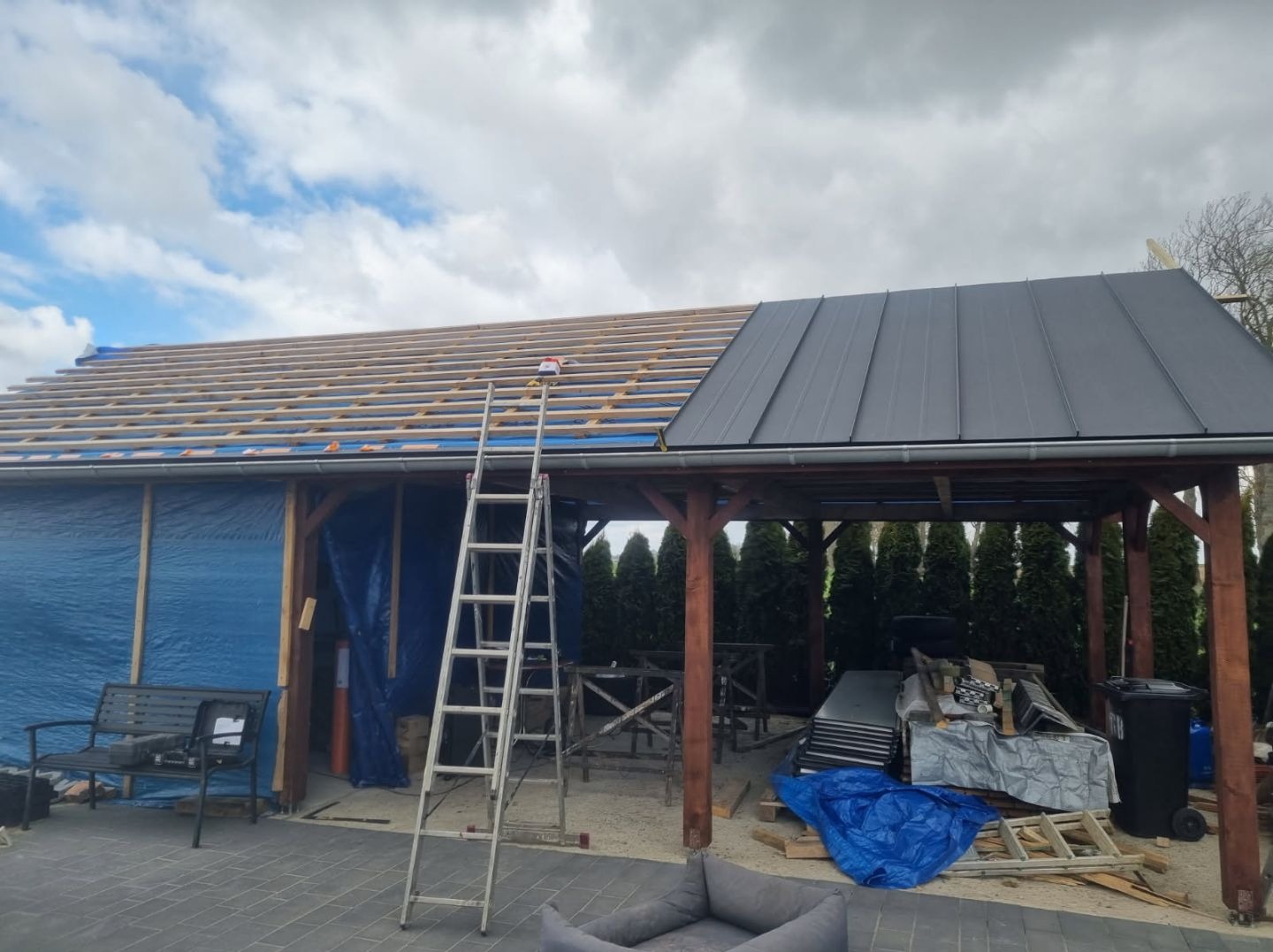 Częściowo pokryty blachą dach drewnianej konstrukcji, z widoczną konstrukcją więźby i drabiną opartą o budynek.