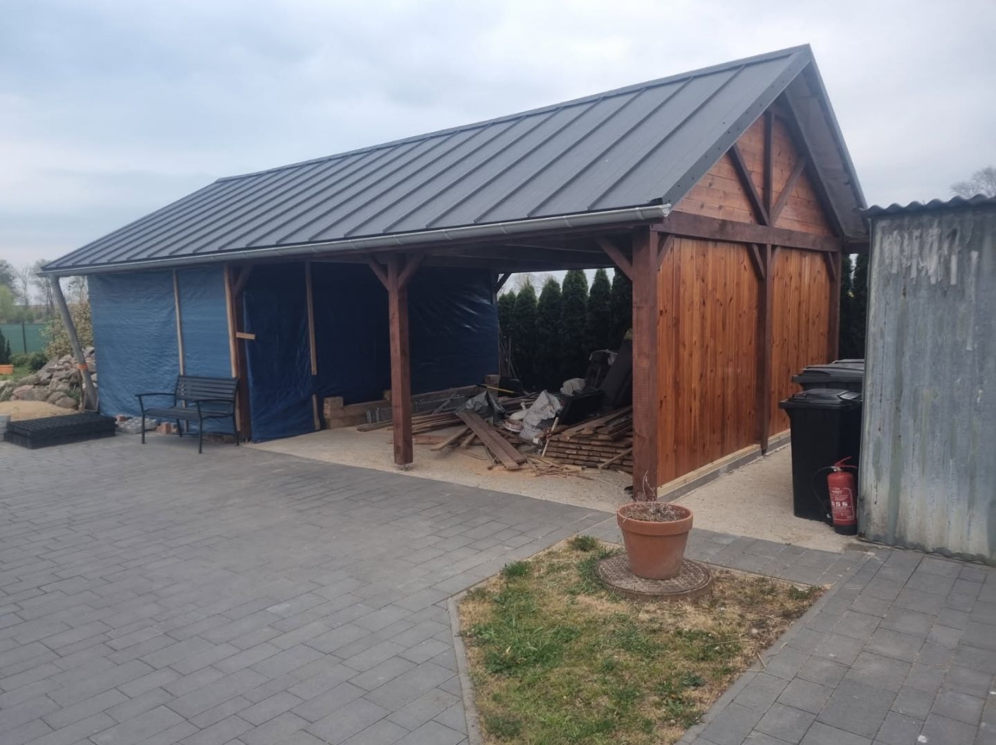 Drewniana wiata z metalowym dachem w trakcie budowy, częściowo osłonięta niebieską plandeką, widoczne deski i narzędzia budowlane, ujęcie z zewnątrz na tle szarej kostki brukowej.