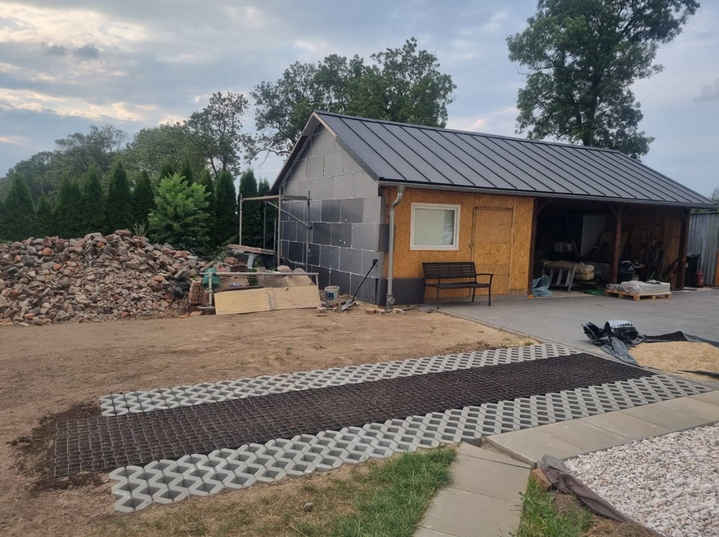 Budynek gospodarczy w trakcie ocieplania elewacji styropianem, z częściowo ułożoną ażurową nawierzchnią z kostki betonowej i stertą cegieł w tle.