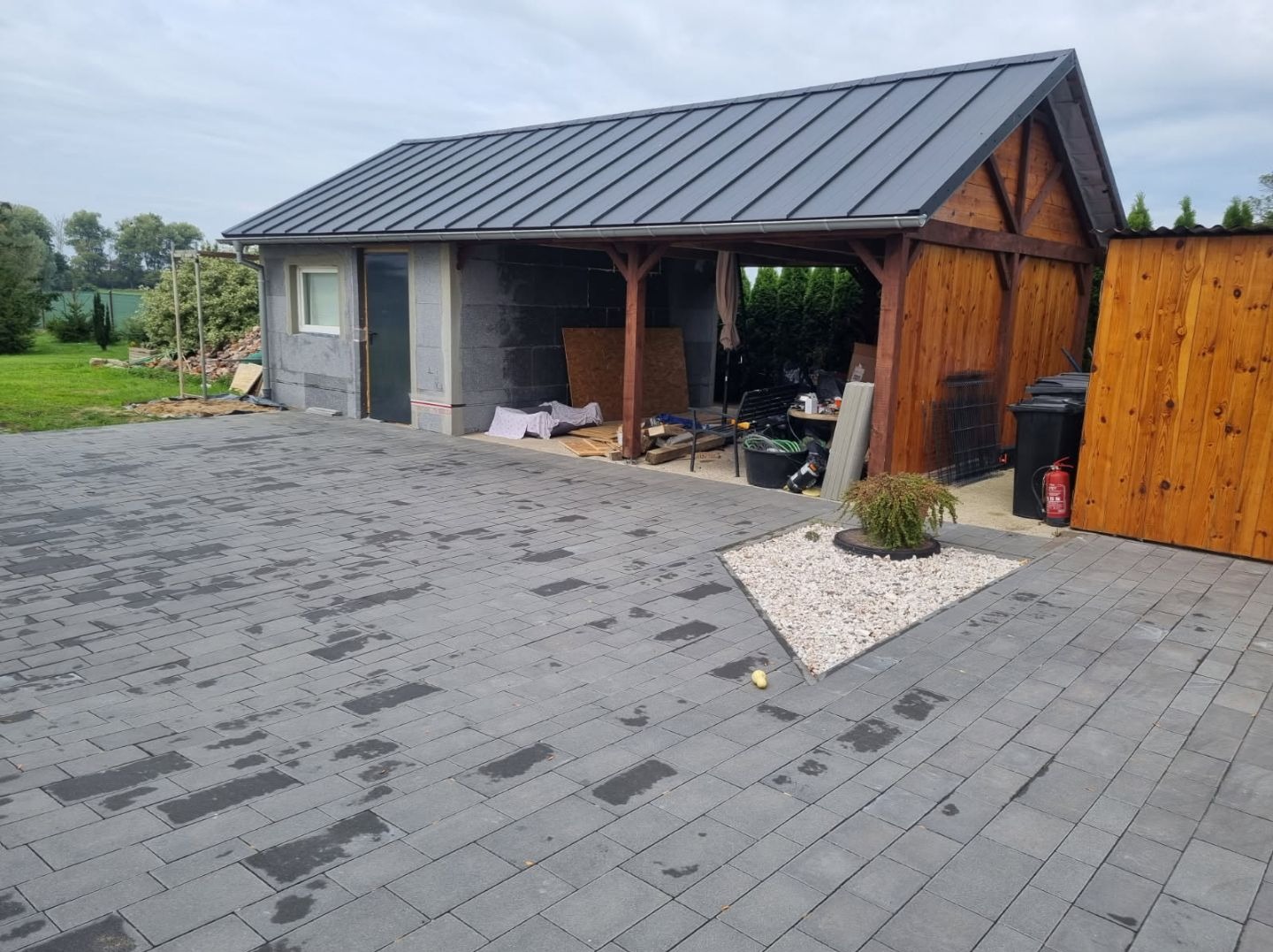 Budynek gospodarczy w trakcie budowy z szarych bloczków i drewnianą wiatą, widoczny fragment podjazdu z kostki brukowej.