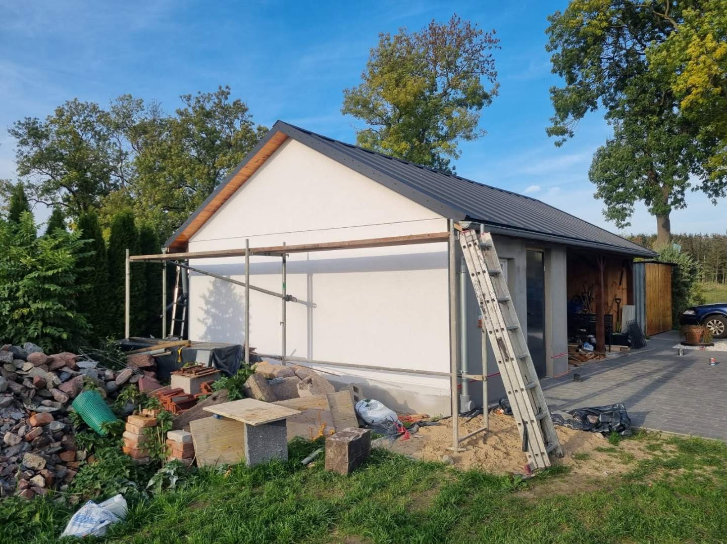 Jednopiętrowy budynek z białymi ścianami, ciemnym dachem i drewnianym wykończeniem szczytu, otoczony rusztowaniem i stertą materiałów budowlanych, w tle drzewa i niebieskie niebo.