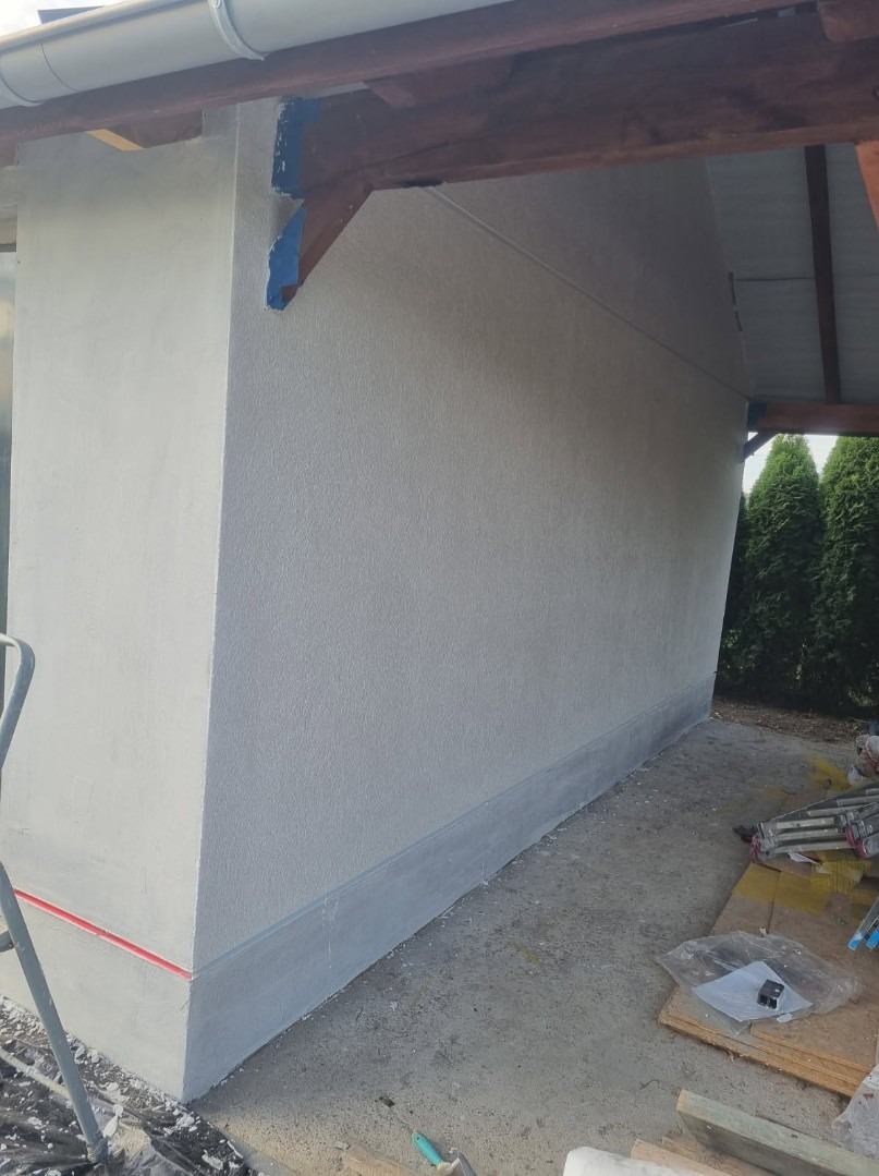 Świeżo pomalowana elewacja budynku z widocznym fragmentem drewnianej konstrukcji dachu i narzędziami malarskimi na podłożu.