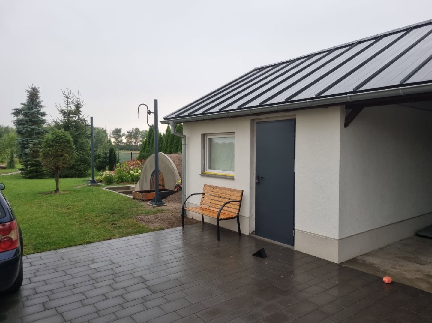 Biały budynek gospodarczy z szarą metalową dachówką, ciemnoszarymi drzwiami, małym oknem i drewnianą ławką przed wejściem, na mokrym brukowanym podjeździe. W tle trawnik, drzewa i element małej...