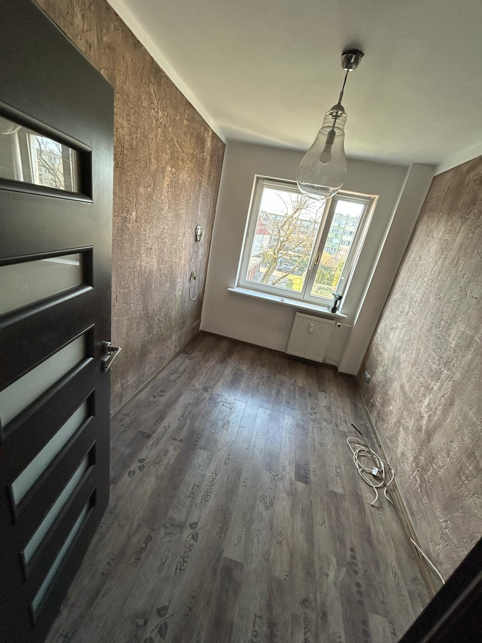 Pokój po remoncie: brązowe ściany o fakturze, drewniana podłoga z nadrukiem, okno z widokiem na drzewo, lampa w kształcie żarówki. Widok zza otwartych, ciemnych drzwi.