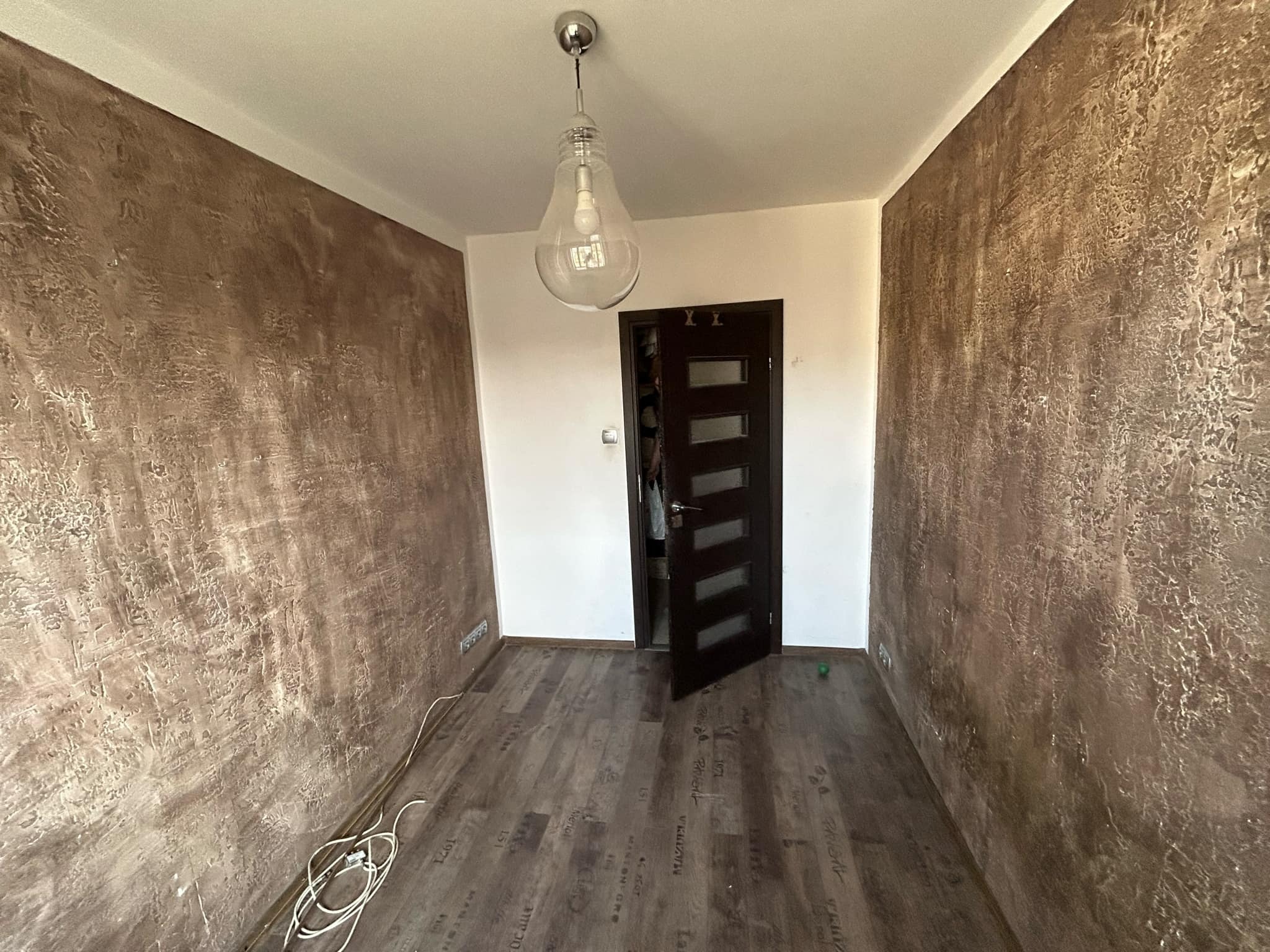 Pokój po wykończeniu ścian z dekoracyjną strukturą w odcieniach brązu, widoczna lampa w kształcie żarówki i drewnopodobna podłoga.