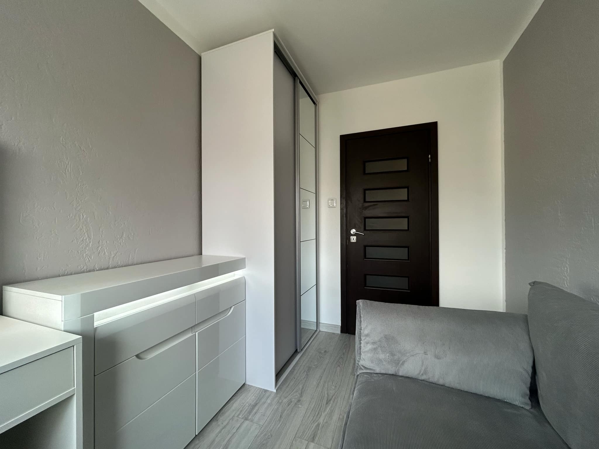 Nowoczesne wnętrze pokoju w szarościach: komoda z oświetleniem LED, szafa przesuwna z lustrem i ciemne drzwi z przeszkleniami, tworzące spójną aranżację.