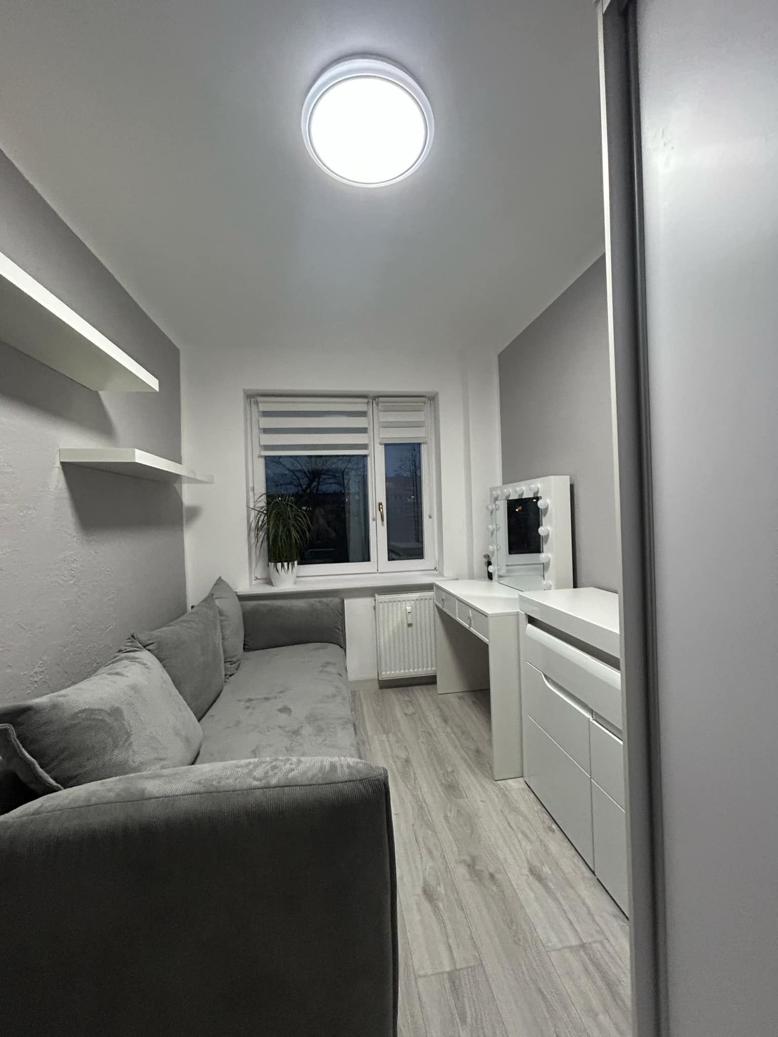 Jasne wnętrze pokoju w odcieniach szarości z sofą, biurkiem z lustrem do makijażu i półkami. Widoczne okno z roletami i lampa sufitowa.