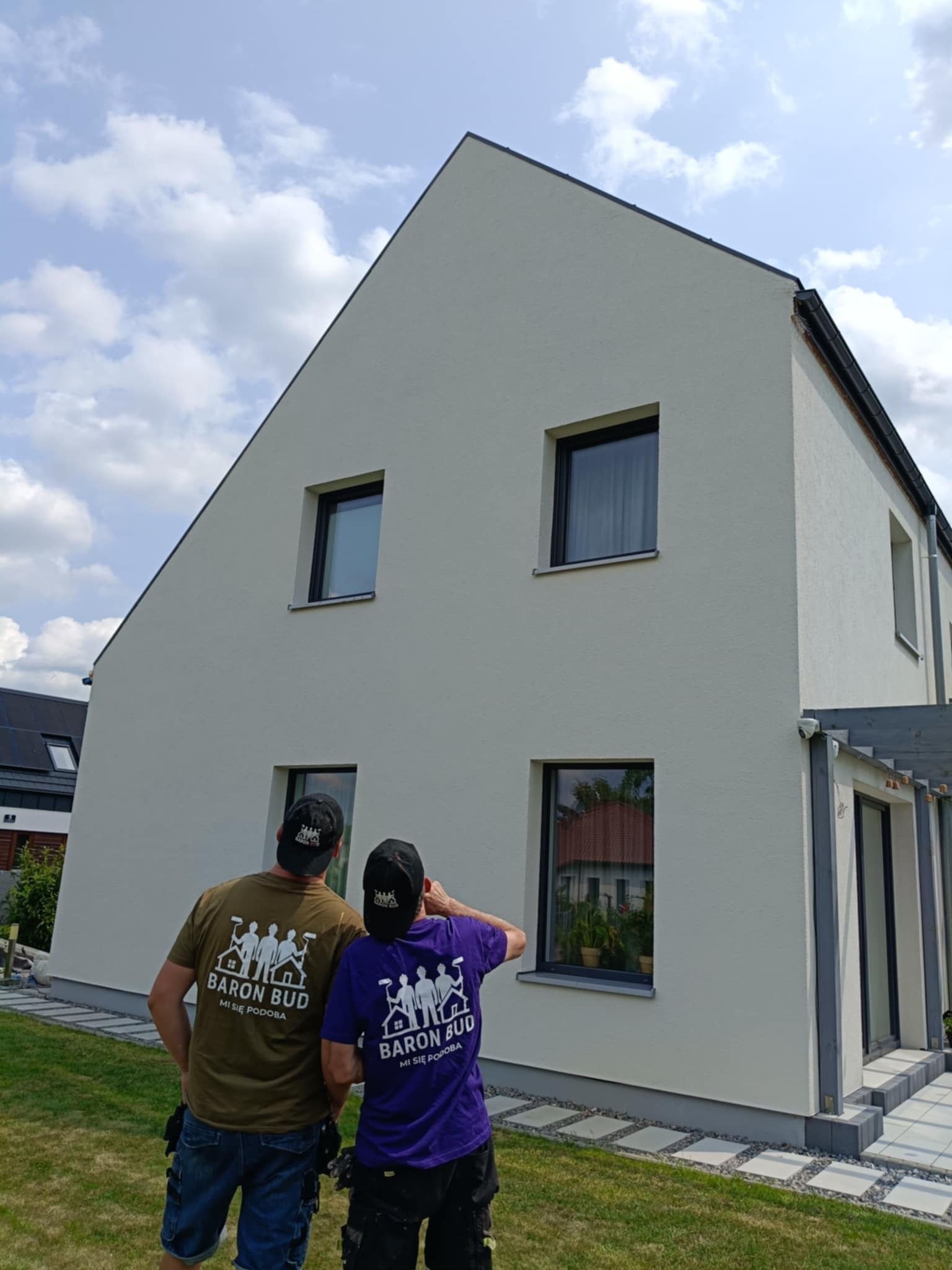 Otynkowana ściana domu jednorodzinnego w jasnym kolorze, z czarnymi ramami okiennymi. Dwóch mężczyzn w koszulkach firmowych przygląda się elewacji.