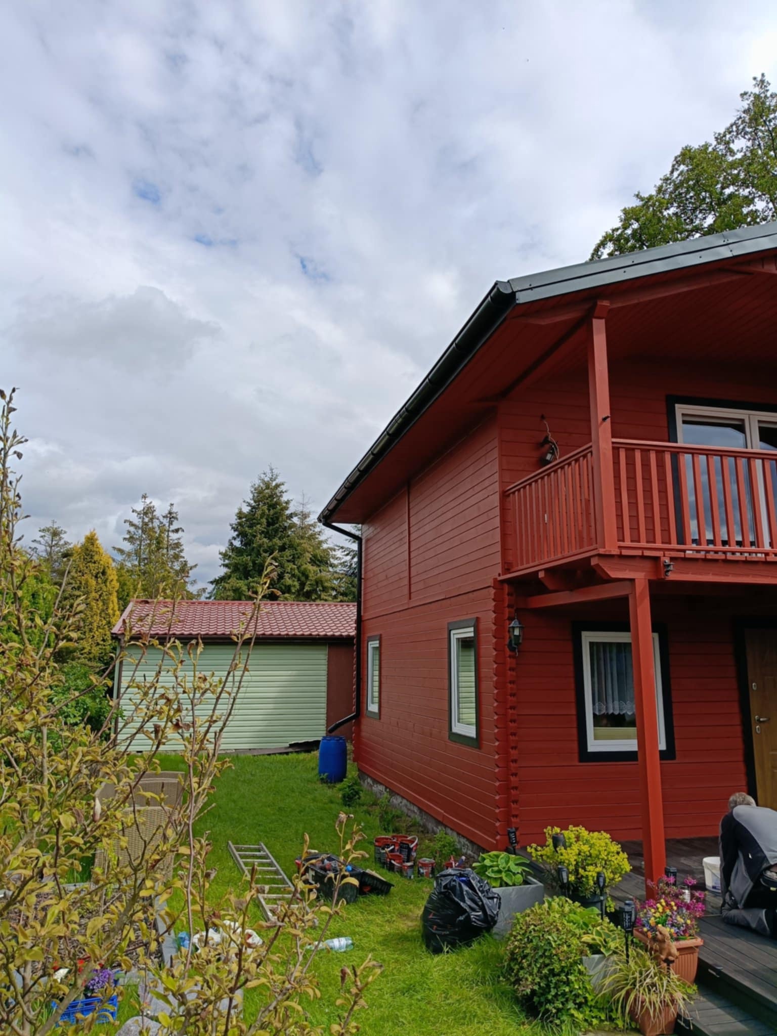 Czerwony, drewniany dom z balkonem w trakcie malowania. Widoczne pędzle, farby i drabina. Obok zielony budynek gospodarczy i osoba pracująca przy elewacji.