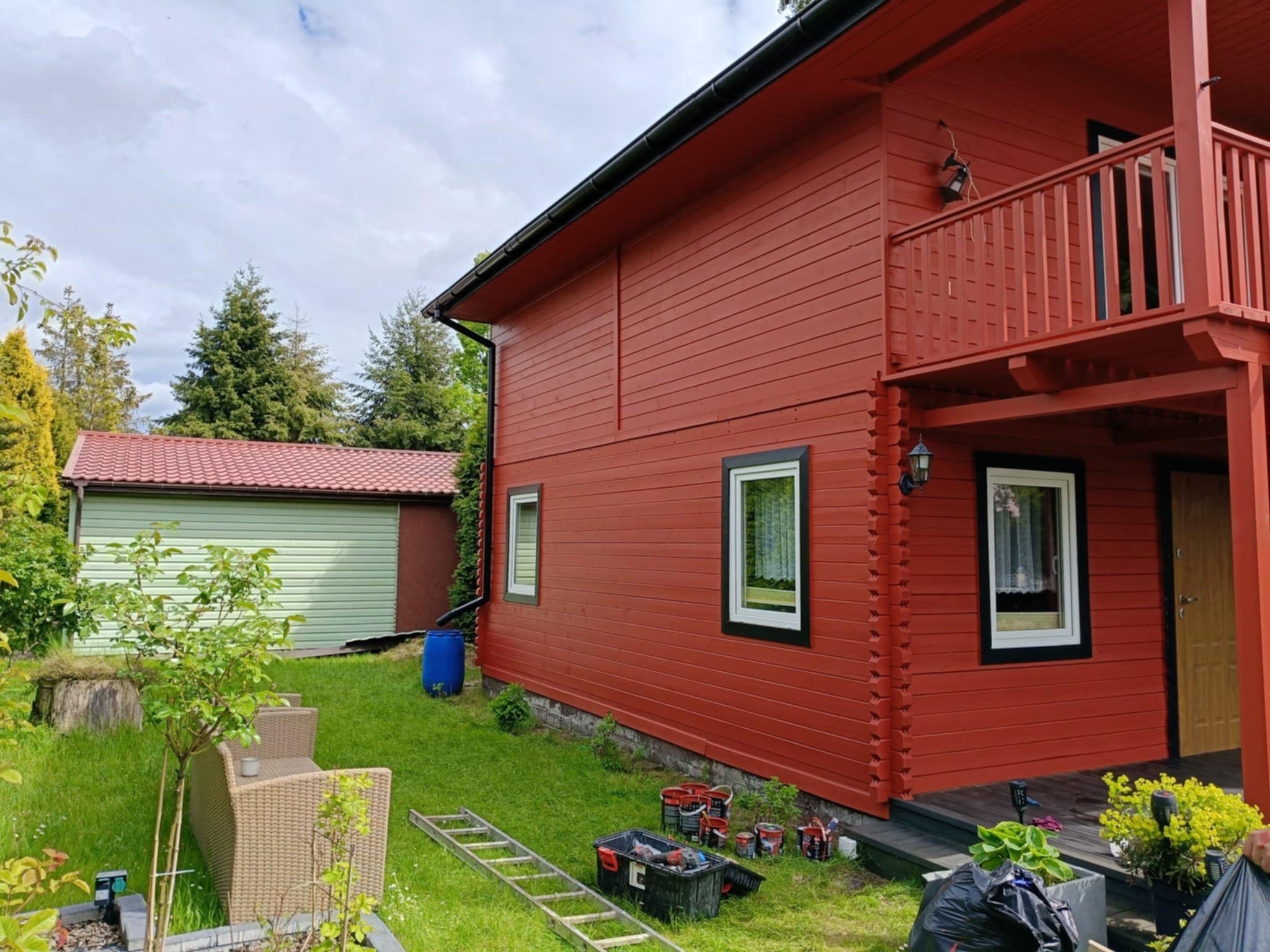 Czerwony, drewniany dom po malowaniu elewacji. Widoczne narzędzia, drabina i puszki z farbą. Trawa, drzewa i budynek gospodarczy w tle.