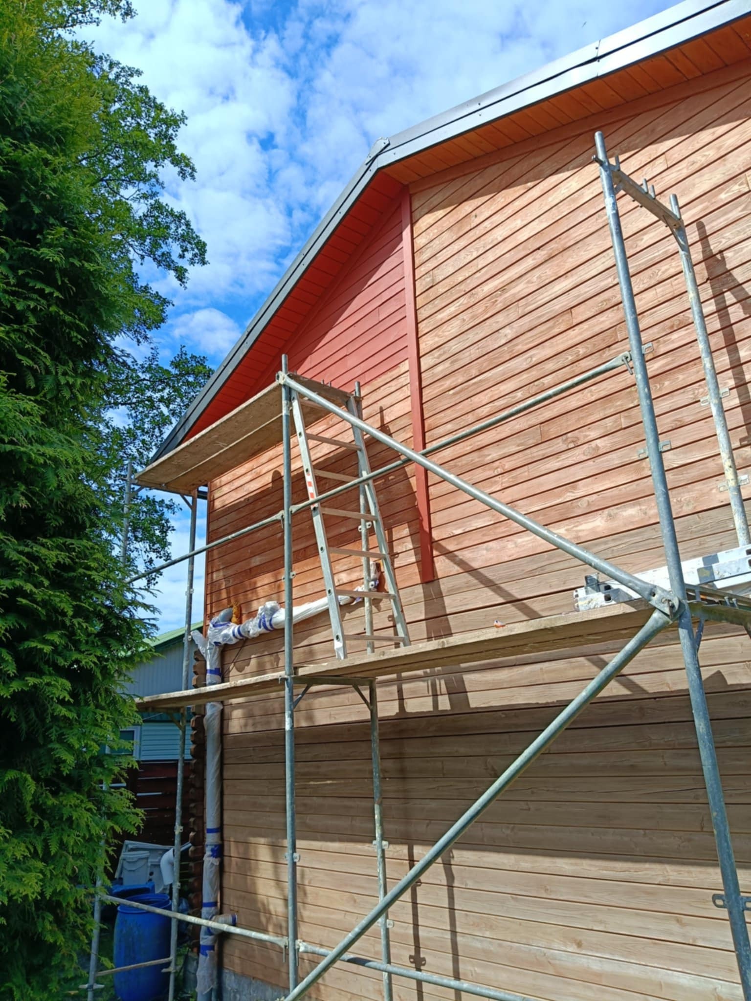 Drewniana elewacja domu w Stargardzie, częściowo pomalowana na czerwono, widoczna z rusztowania. Świeże malowanie, błękitne niebo w tle.