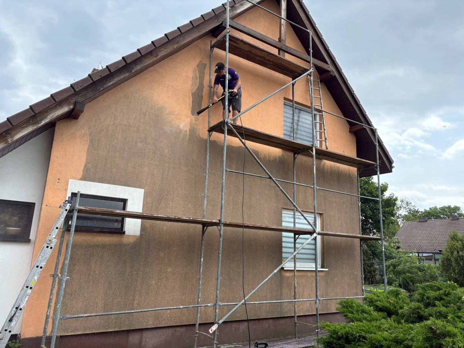 Pracownik na rusztowaniu czyści elewację domu myjką ciśnieniową. Widoczny fragment wyczyszczonej elewacji o jaśniejszym odcieniu. Na pierwszym planie metalowe rusztowanie.