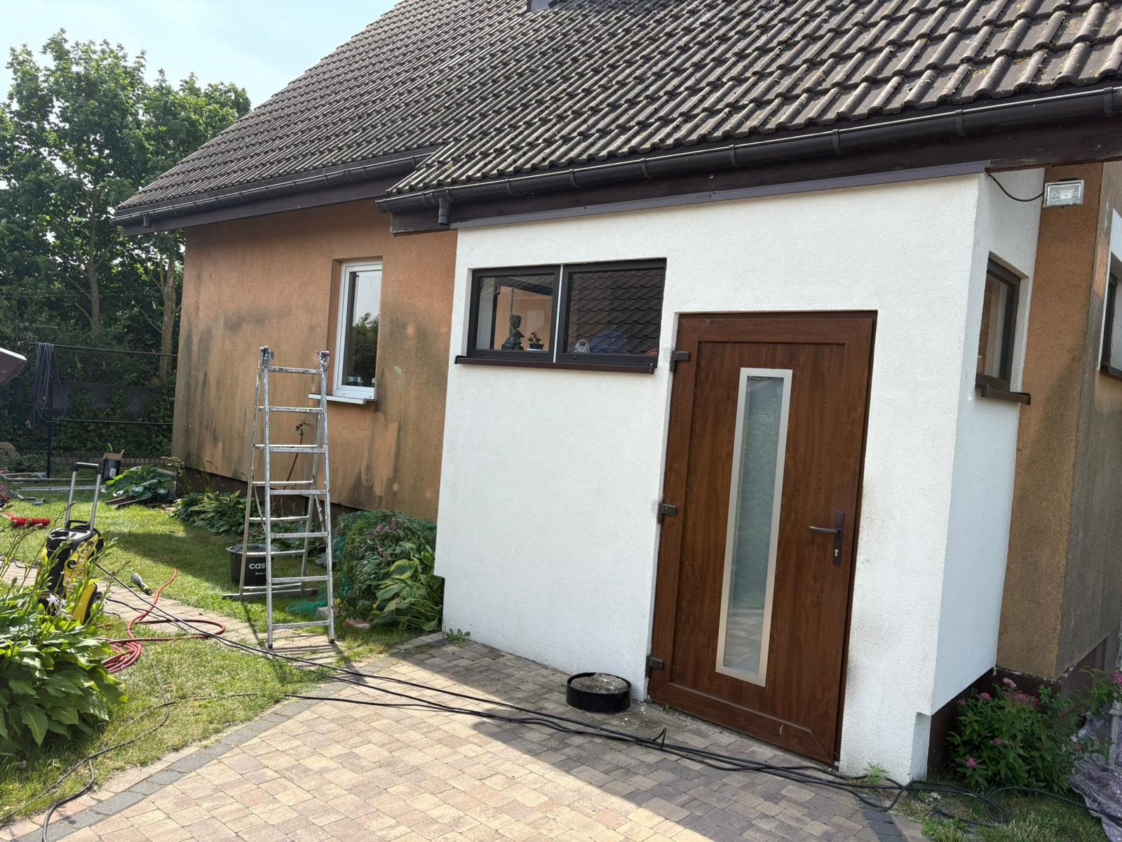 Elewacja domu przed i po czyszczeniu: część brązowa, część biała. Widoczna drabina i myjka ciśnieniowa. Brukowany chodnik i zieleń wokół.