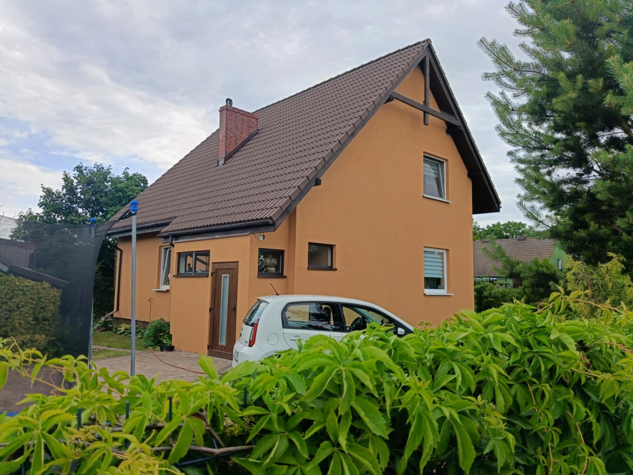Dwukondygnacyjny dom z pomarańczową elewacją i brązowym dachem, częściowo zasłonięty zielenią, z białym samochodem na podjeździe, w pochmurny dzień.