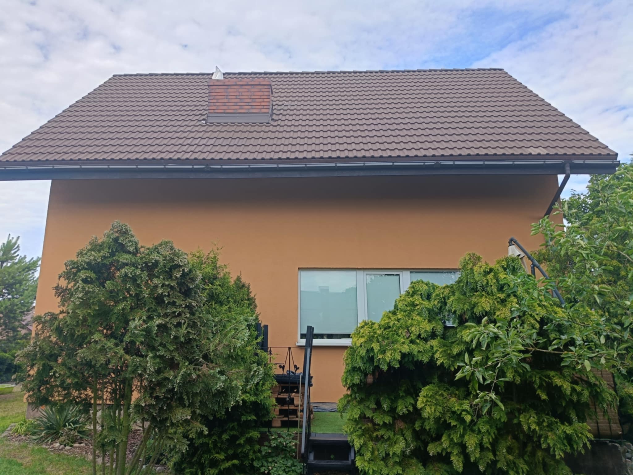 Dom z brązowym dachem i pomarańczową elewacją widziany z poziomu ogrodu, częściowo zasłonięty przez zielone krzewy.