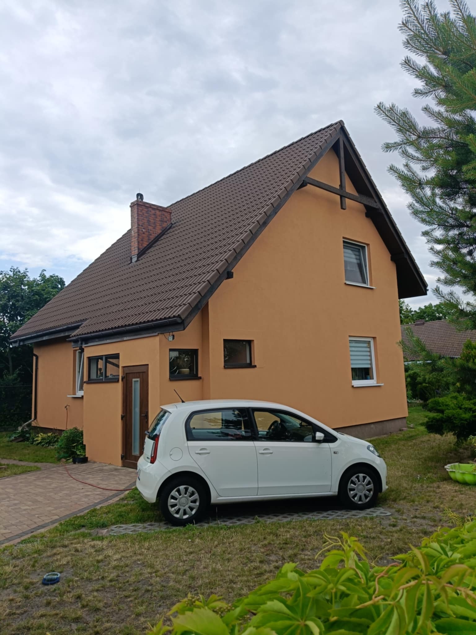 Dom jednorodzinny z pomarańczową elewacją i brązowym dachem, obok stoi biały samochód. Widoczne elementy wykończenia, takie jak okna i drzwi. Ujęcie z poziomu gruntu.